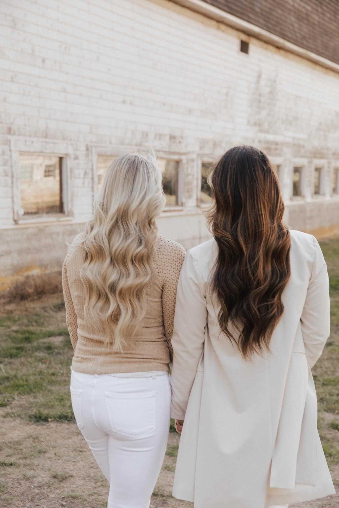 Two women walking together along a white building with rustic windows, seen from behind.