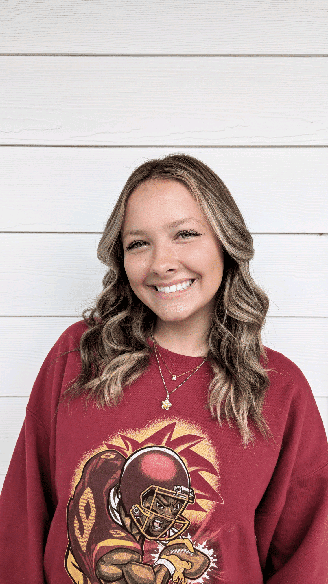 Girl in a red football-themed sweatshirt smiling against a white wooden wall.