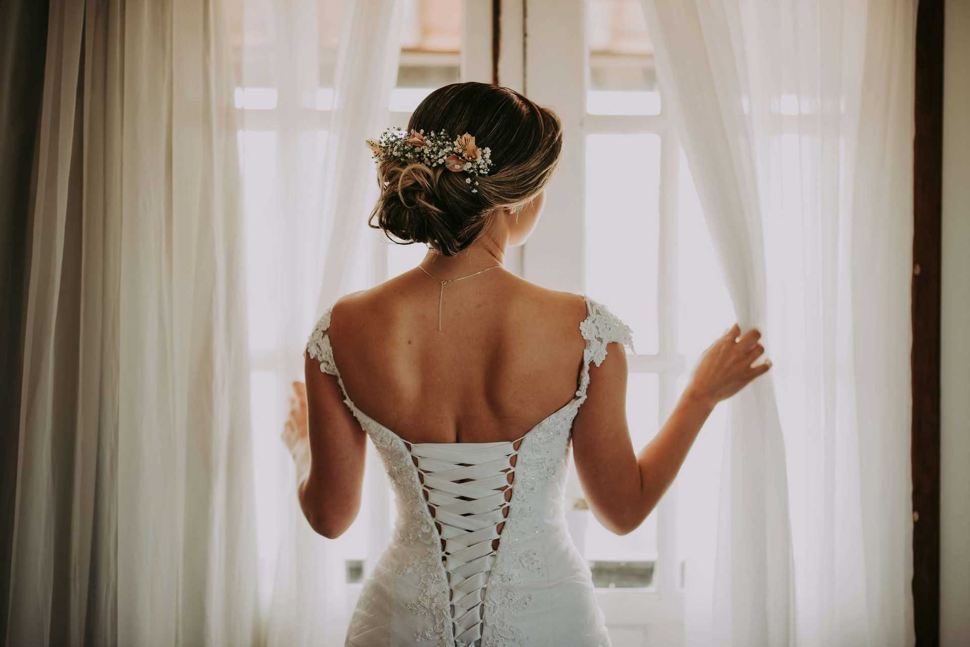 Bride in elegant lace dress gazes out window, soft light illuminating her floral hairpiece.