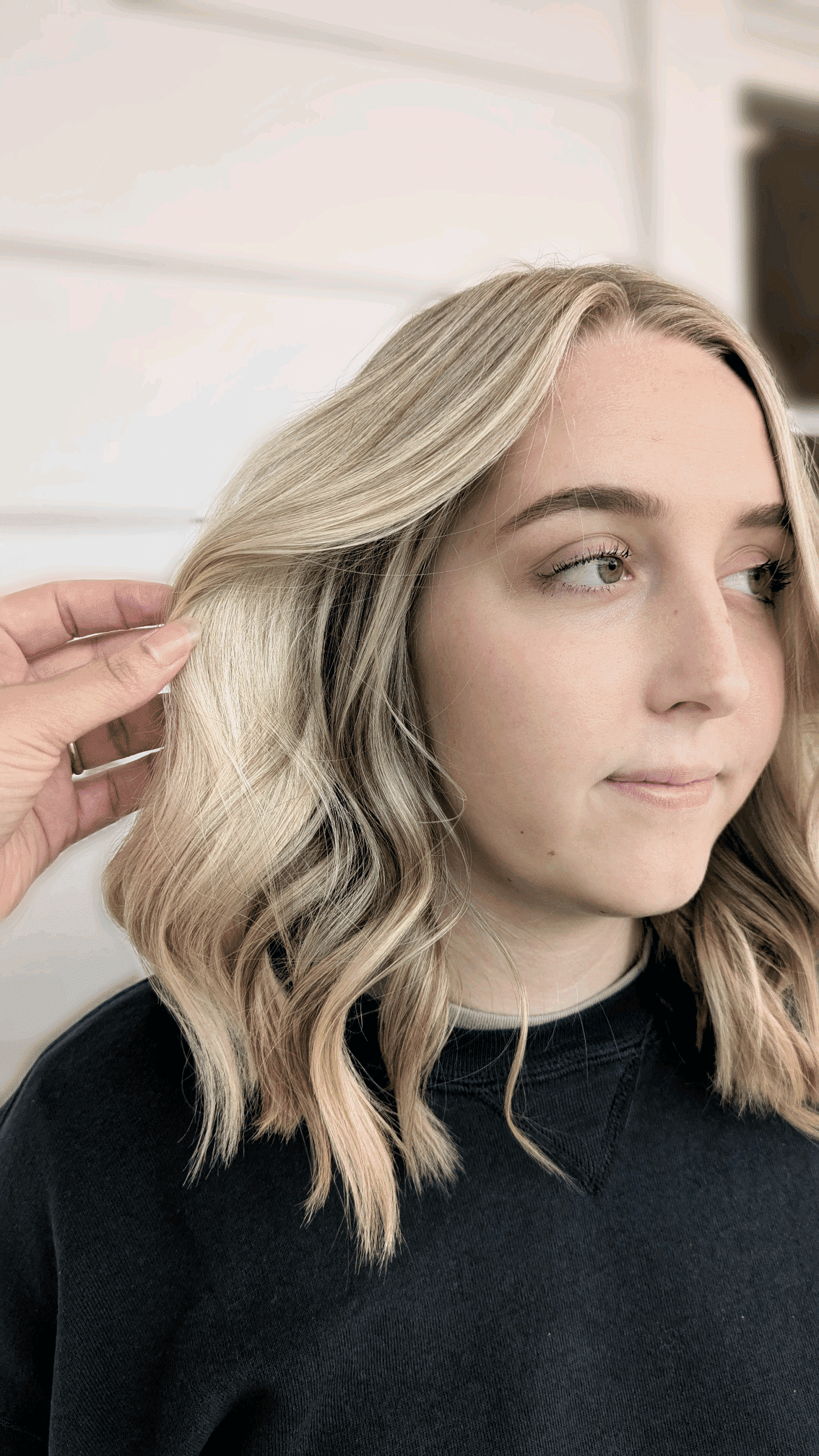 Person with wavy blonde hair styled; hand adjusting a curl, soft background.