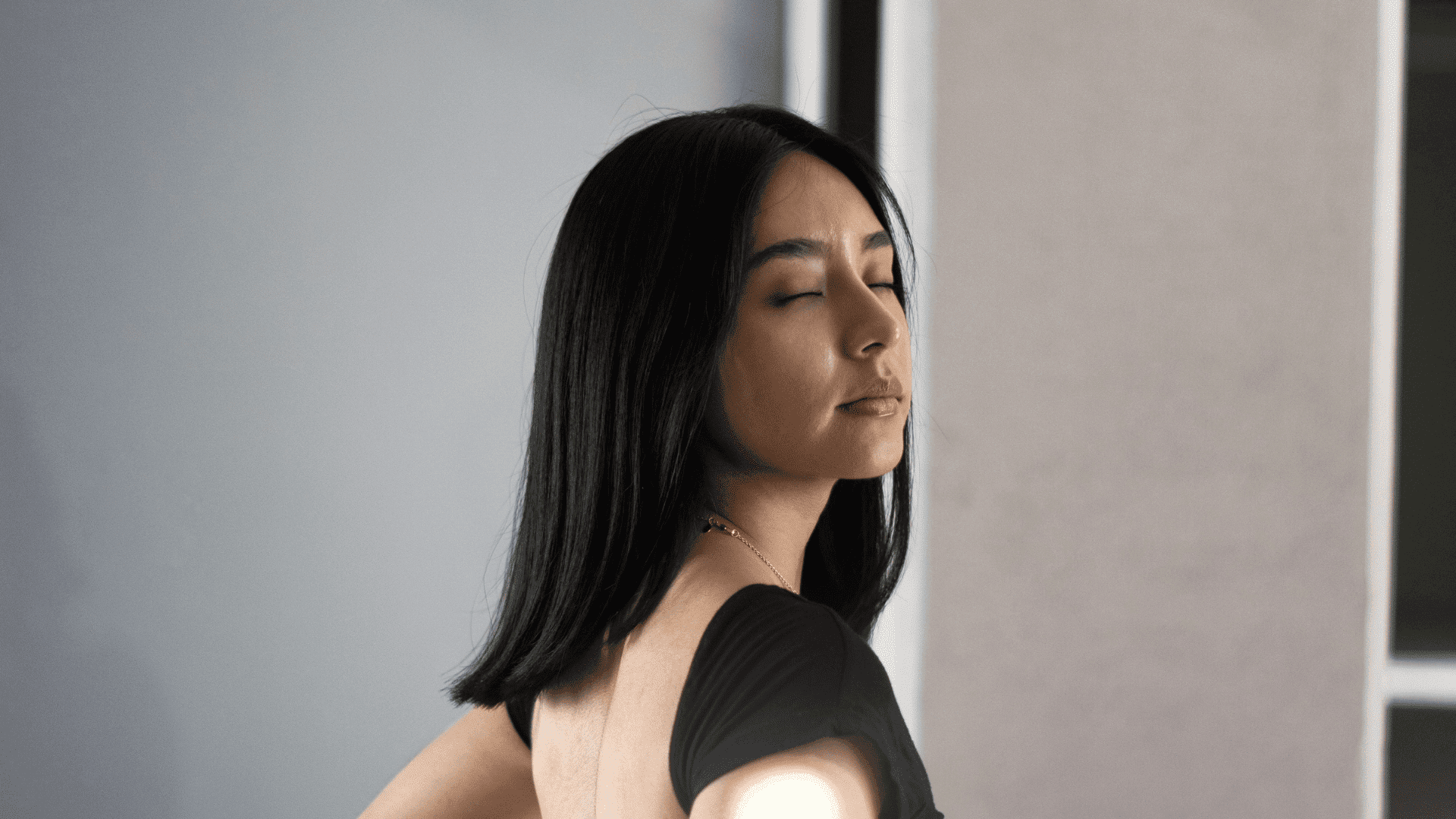 Woman with closed eyes in a black top, standing against a neutral background.